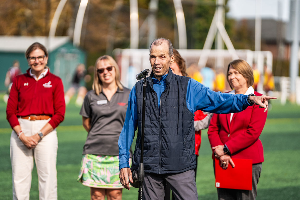 A man in a vest speaks to a crowd on a soccer field